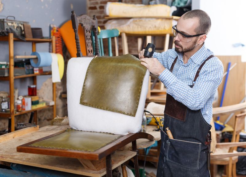 Craftsman upholstering a chair seat with green leather in a furniture workshop.