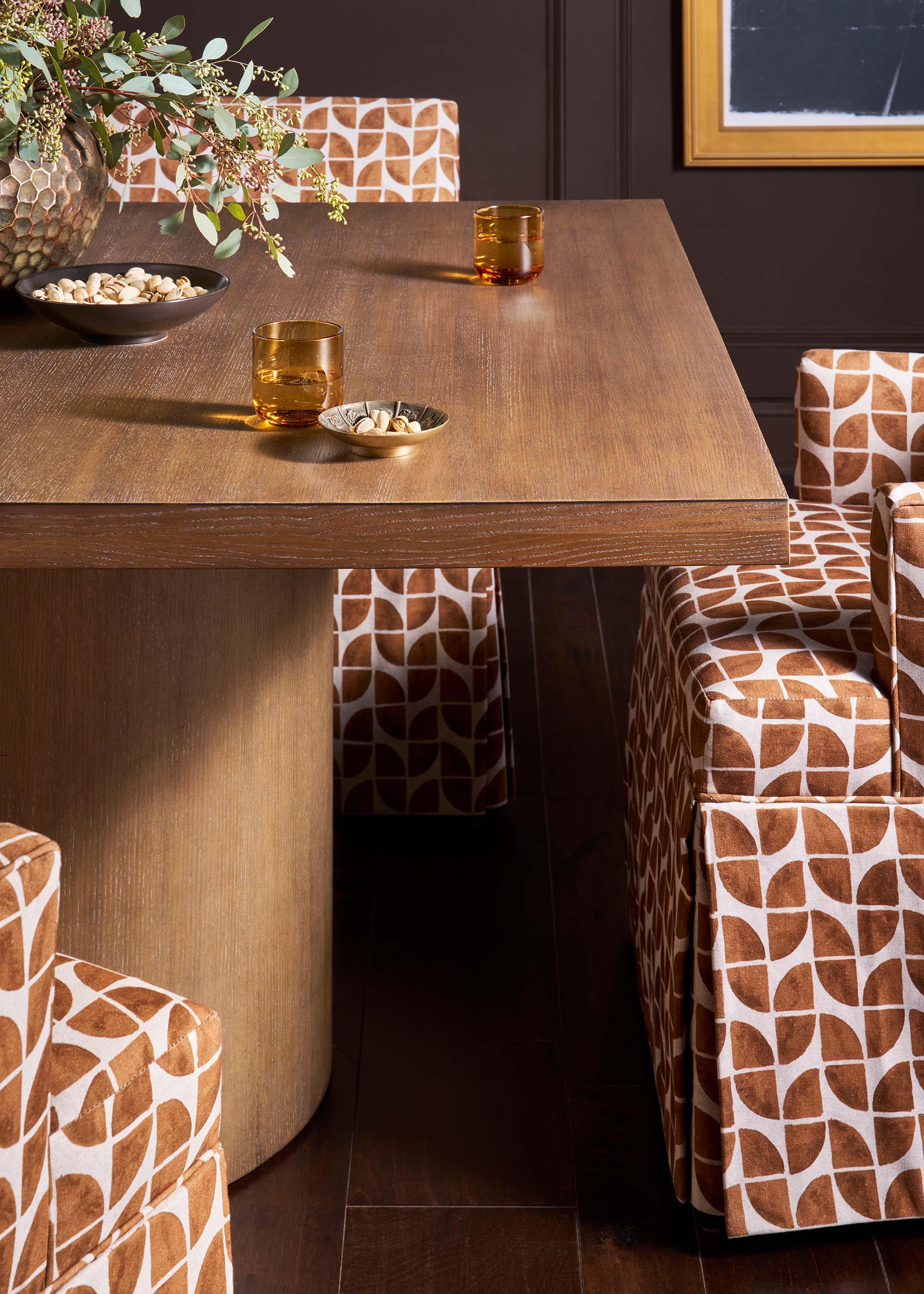 Close-up of a wood dining table with a textured walnut finish, surrounded by chairs upholstered in a rust and white geometric print, with an amber glass and small dish on the tabletop.