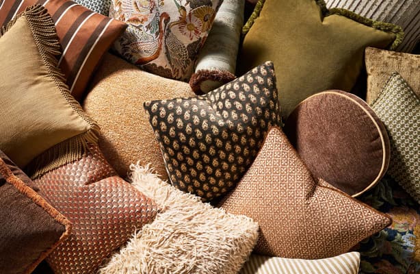 Pile of throw pillows in warm brown, rust, olive, and cream tones, mixing patterns such as stripes, paisley, diamond prints, and fringed and shaggy textures.