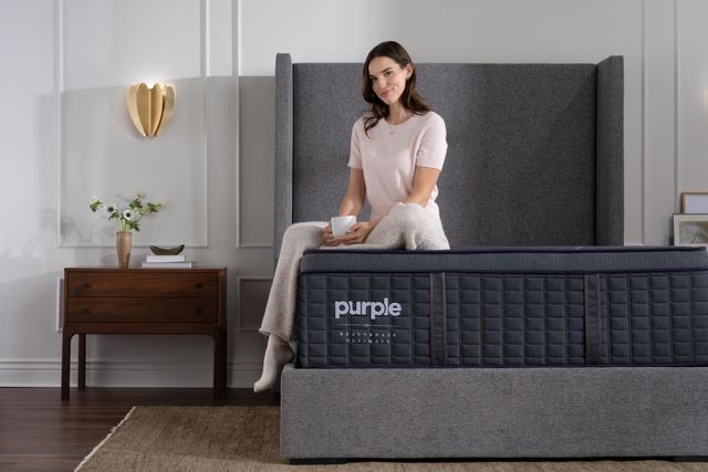 Front view of a woman sitting and smiling on a Purple mattress in a modern grey bedroom.