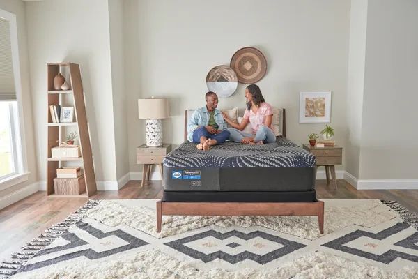 Front view of a casual bedroom with a couple sitting and smiling on a Sealy Posturepedic mattress
