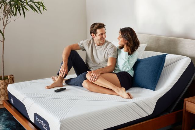 Front view of a couple sitting together and smiling on a mattress atop an adjustable base