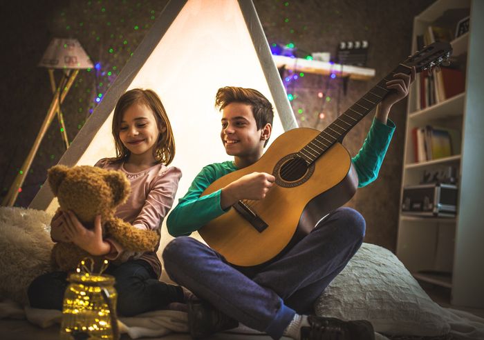 young brother and sister play the guitar together while sitting outside their indoor tent