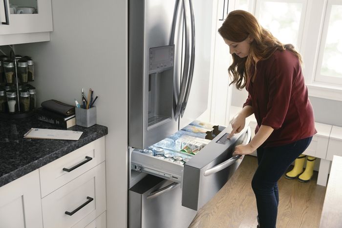 A woman stands in front of a Frigidaire 4-Door refrigerator, pulling open the Custom-Flex Temp Drawer.