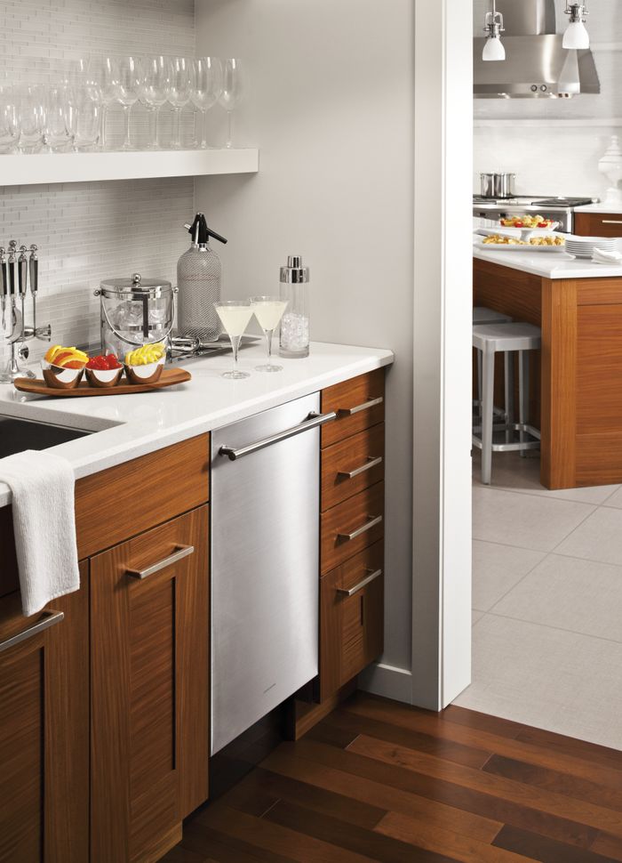 A clean, white mini bar leading into a kitchen, with white walls and brown wooden cabinets. A dishwasher is built into the cabinets with cocktail fixings on the counter.