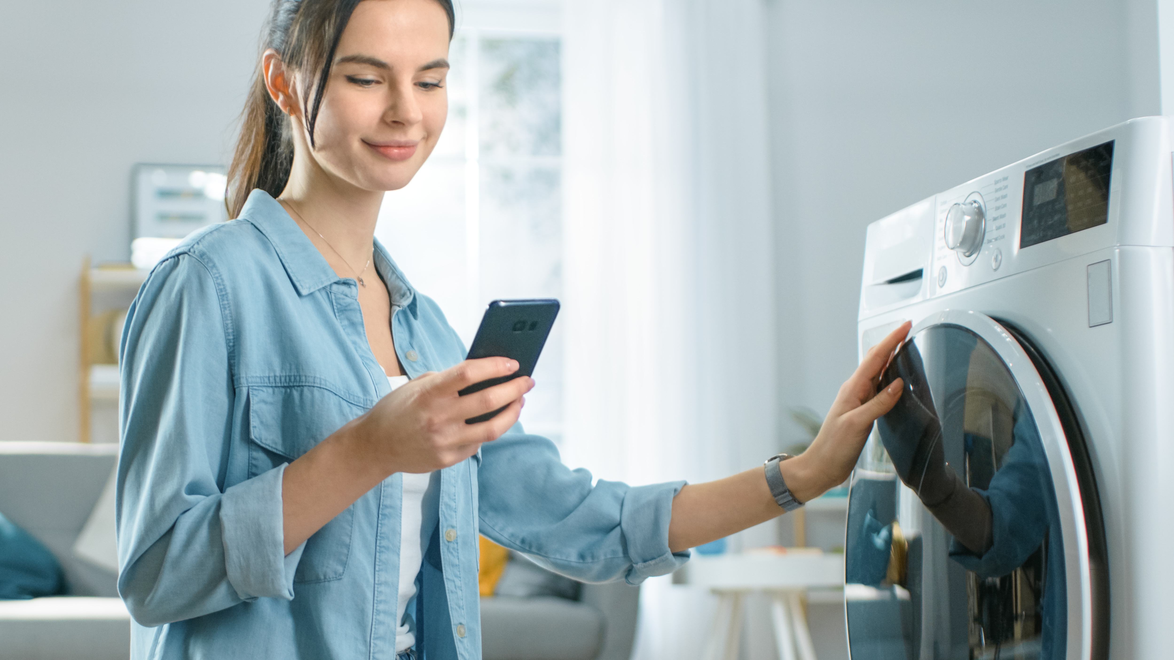 a young woman uses her smartphone to set the cycle on her washer