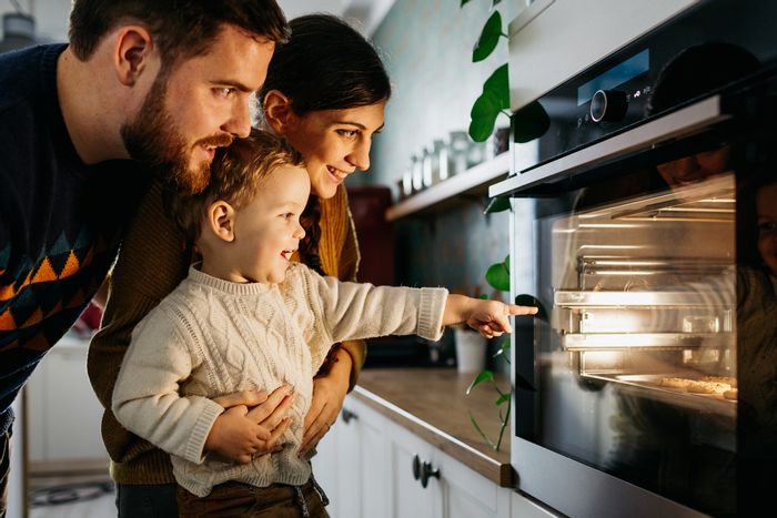 A baby and his parents look into a wall oven at their baking cookies