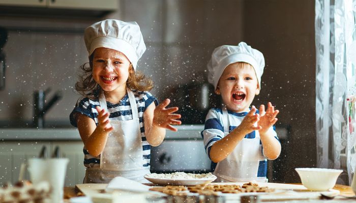 Two siblings in matching shirts, aprons and chefs hats clap flour across their cookie workstation 