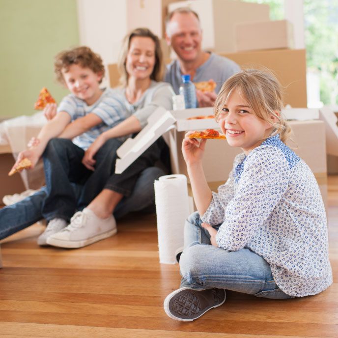 A family sitting on the floor of their home eating pizza together, with boxes piled up in the background.