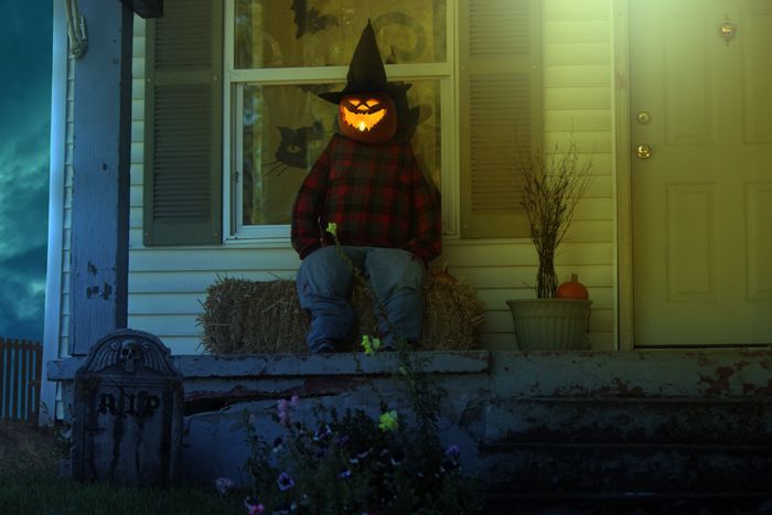 Scarecrow with jack o' lantern head and witch hat sits outside of a dark house.