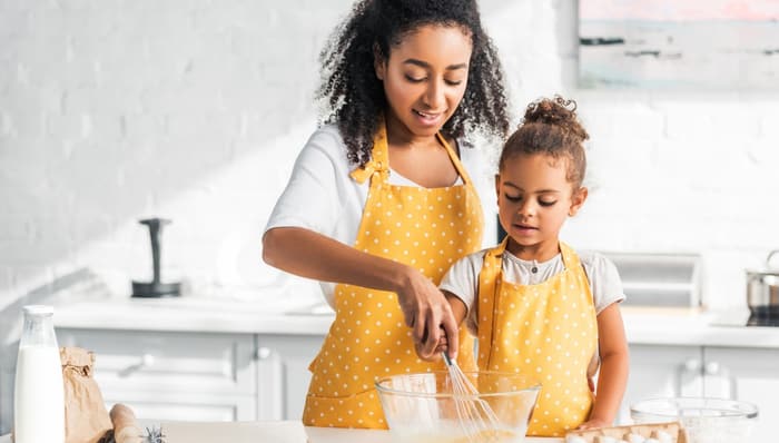 A young mother and her daughter mix a bowl of eggs together in a white kitchen.