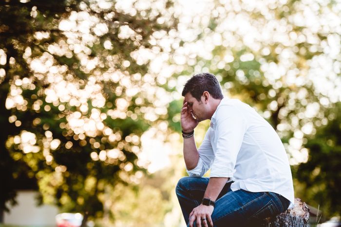 Man sitting on a tree stump with trees in the background. He looks tired, with his head in his hands.