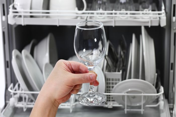 A hand holds up a perfectly clean wine glass in front of an open dishwasher full of clean dishware.