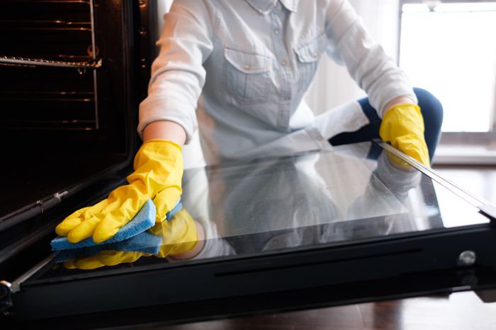 A woman scrubs the inside of an oven door in a white kitchen.
