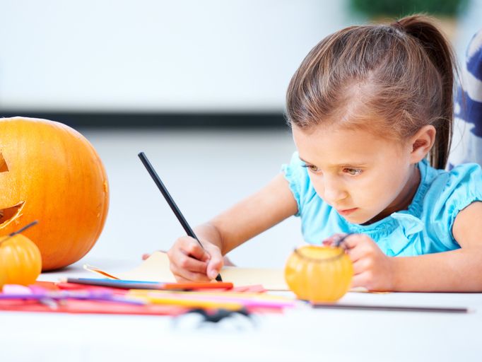 Little girl sitting at a table and coloring. A jack o' lantern is on the table beside her.