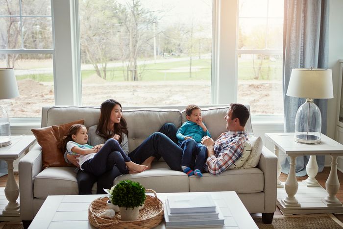 A couple and two young children lounge on a beige couch in a traditional-styled living room with a window open to their front yard.