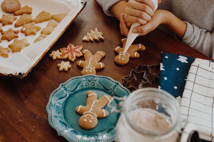 Freshly made gingerbread men lay scattered on a table while a pair of hands work at icing one cookie with a piping bag in the upper right corner.