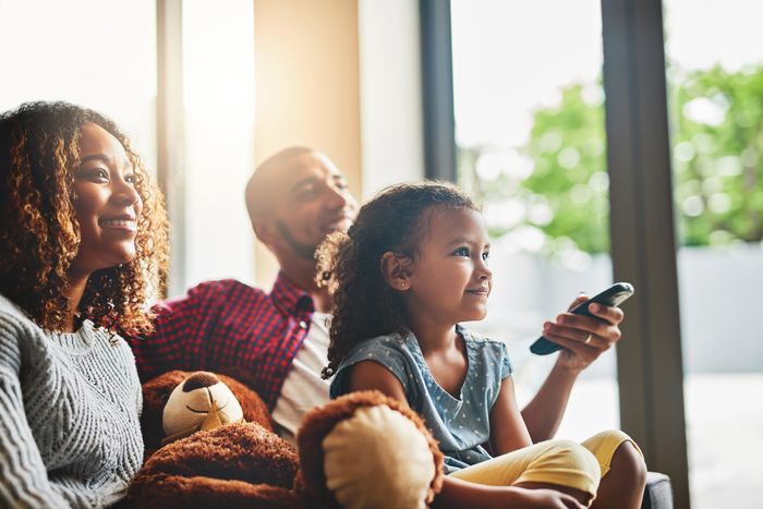 A mother, father, and young daughter sit together on the couch. The father holds a television remote.