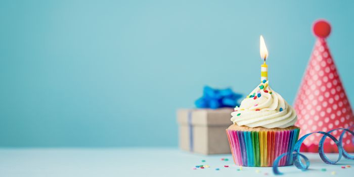 A vanilla cupcake with a single, yellow candle stuck into the frosting sits on a table in front of a blue backdrop. A small gift box and a pink party hat are in the background.