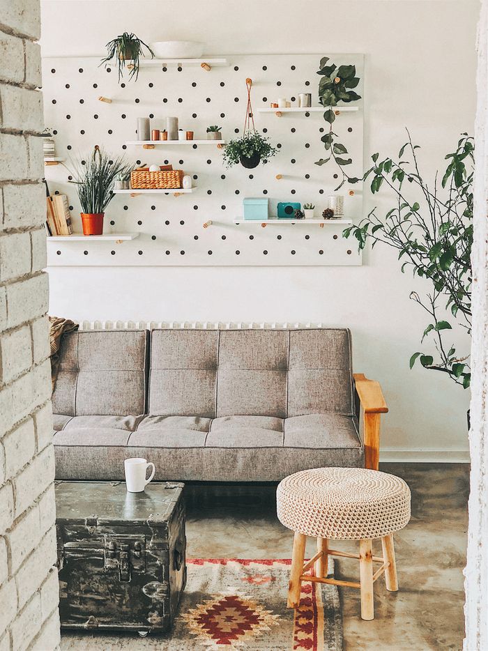 A grey futon in a white apartment. A pegboard hangs on the wall above it, covered in shelves holding books and hanging plants.