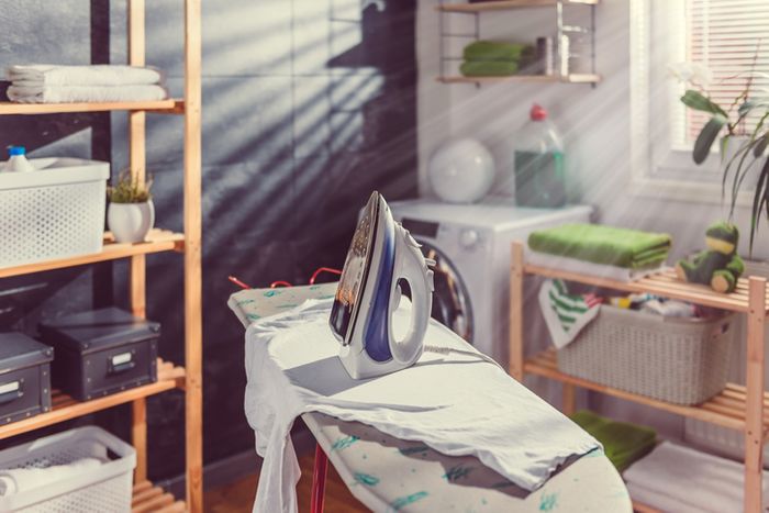Clothes iron standing up on an ironing board in a sunny laundry room. Baskets of clothes and a laundry machine can be seen in the background.