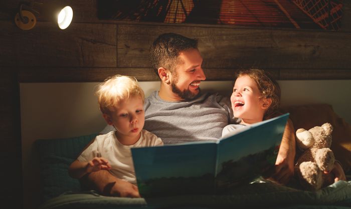A young father sits in bed with his two young children, reading a picture book.