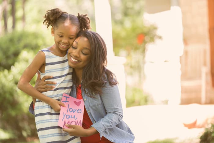A mother and daughter embrace. the daughter is holding a handmade card that reads "I love you, mom"