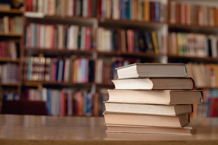 A stack of hardcover books sit on a wooden table, with shelves full of books visible in the background.