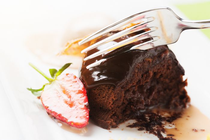 Slice of chocolate cake on a white plate with half a strawberry on it. A fork is being used to cut away a piece.