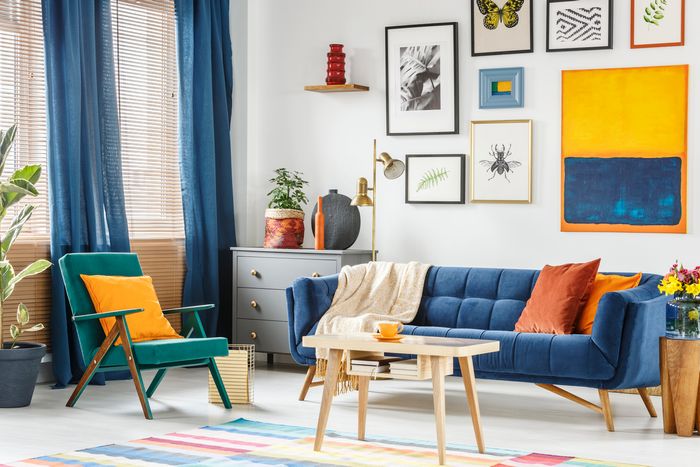 A bright, white living room with a deep blue couch, blue drapes, and orange and yellow accent pillows.