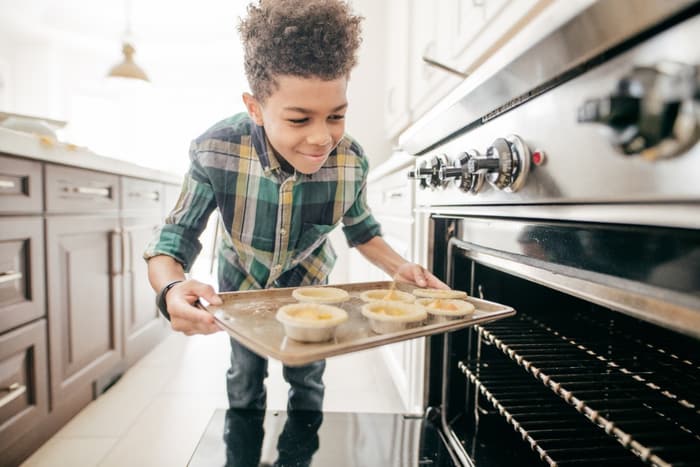 A young boy puts a tray full of quiches into the oven.