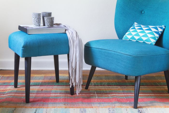 A blue upholstered chair with a small, square matching ottoman with tall legs made of dark wood. A book, a scarf, and three candle holders are stacked on top.