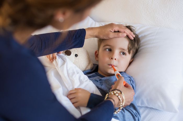 Child in bed with a thermometer in his mouth and a woman sitting at the edge of the bed taking his temperature.