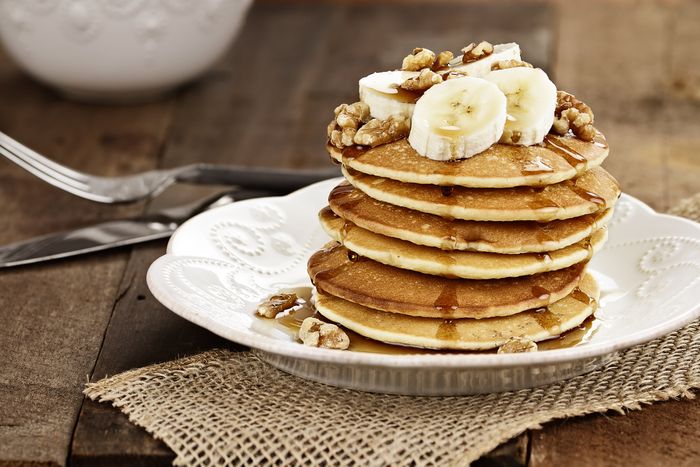A stack of pancakes sits in a fancy white dish, on a rustic, wooden table. The cakes are topped with sliced bananas, walnuts, and syrup.