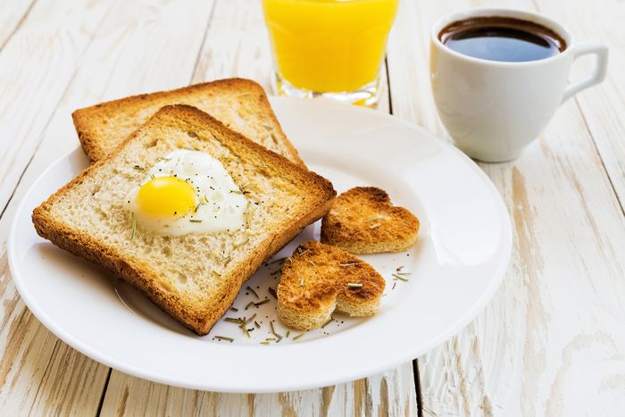 Two pieces of toast on a plate, alongside cups of coffee and orange juice. Heart chaped holes are cut out of each and a fried egg is in each of the holes.