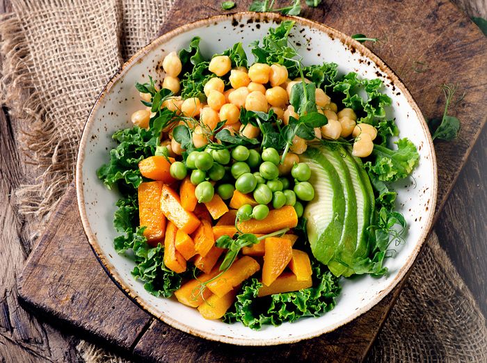 A white bowl full of kale, squash, chickpeas, avocado, and beans sits on a rustic, wooden table.