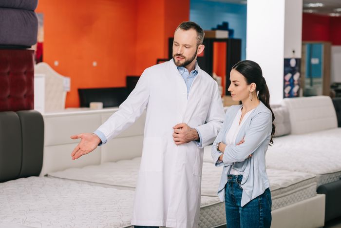 A woman shopping for mattresses stands next to a man in a white coat, who shows her different options.