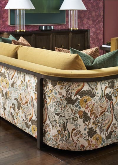 Close-up of the back and side of a sofa upholstered in a cream fabric printed with cranes, florals, and foliage, topped with mustard-yellow cushions and patterned throw pillows, set against a dark wood credenza and patterned wallpaper.