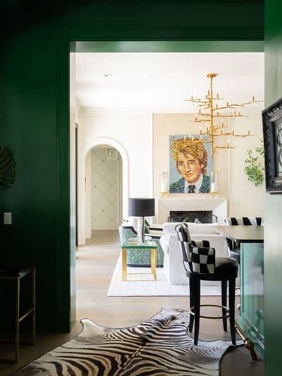 View through a green-framed doorway into a living room with a mosaic portrait above a fireplace, a branch-style gold chandelier, a white sofa, a green patterned armchair, and a checkered-upholstered barstool, with a zebra-print rug in the foreground.