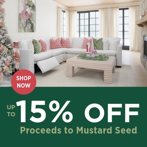 A holiday-themed living room with a white sectional sofa decorated with pink and green patterned throw pillows. A light wood coffee table sits in front of the sofa with festive greenery and small Christmas figurines on top. A decorated Christmas tree with pink and gold ornaments is partly visible on the left. On the right side of the image, a dark green banner displays the text UP TO 15% OFF in large white letters, with a red circular button reading SHOP NOW. Smaller text at the bottom reads, Proceeds to Mustard Seed.
