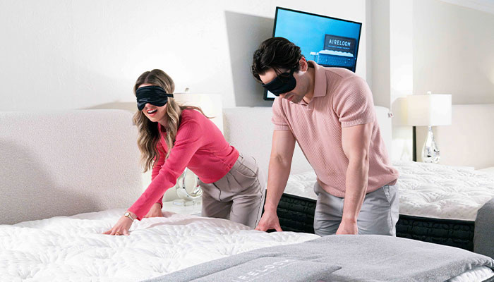 Blindfolded couple testing mattresses in a mattress showroom