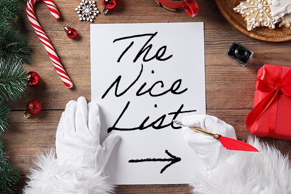 Santa writing The Nice List on a sheet of paper placed on a wooden tabletop surrounded by a candy cane, gift ornament and pine tree.
