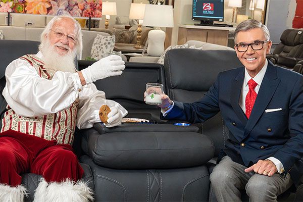 Bob Mills and Santa sitting on a recliners, cheering with a glass of milk and cookies.