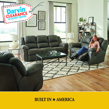 Dining room showroom display featuring a dark wood rectangular table with four matching chairs and a coordinating bench, set on a light patterned area rug. A vase of yellow flowers sits on the table beneath two black pendant lights, with large windows in the background. A “Darvin Clearance & Outlet Center” badge appears in the top left, along with “Price Plunge” and “While Quantities Last!” promotional text.