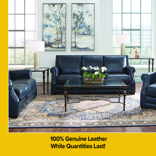 Living room showroom featuring a black leather sofa, matching loveseat, and chair arranged around a white rectangular coffee table with a glass shelf. The seating sits on a light patterned area rug over hardwood floors, with a side table, lamp, tall potted plant, and two framed abstract prints on the wall. Promotional text reads “Price Plunge” and “100% Genuine Leather wherever the body touches. While Quantities Last.”