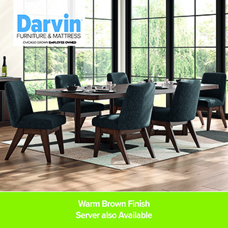 Dining room set with a rectangular wooden table in a warm brown finish surrounded by six upholstered blue chairs, placed on a light rug in a bright room with large grid windows. Darvin Furniture & Mattress branding appears in the corner, with text reading “Warm Brown Finish. Server also Available.”