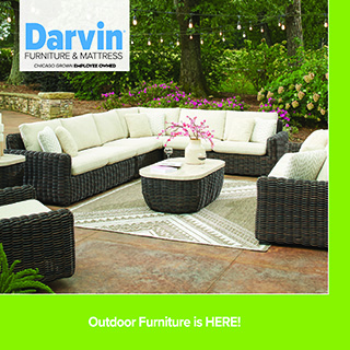utdoor patio seating area with a large sectional wicker sofa and matching chairs arranged around a round coffee table on a patterned outdoor rug, set in a lush garden with trees and flowers. Darvin Furniture & Mattress branding appears, with text reading “Outdoor Furniture is HERE!”