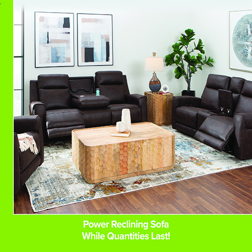 Living room setup featuring a dark brown power reclining sofa and matching loveseat arranged around a rectangular wooden coffee table on a patterned rug. Wall art, a side table with a lamp, and a potted plant decorate the space. Darvin Furniture & Mattress branding appears with text reading “Power Reclining Sofa While Quantities Last!”