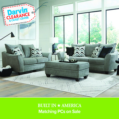 Light gray living room set with a sofa, loveseat, and matching ottoman arranged on a textured rug in a bright room with large windows. A Darvin Clearance & Outlet Center tag appears in the corner, with text reading “Built in America – Matching PCs on Sale.”