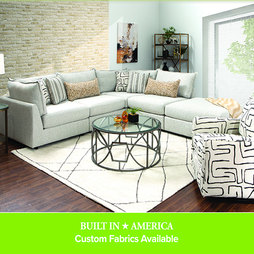 Light gray sectional sofa with patterned accent pillows arranged around a round glass coffee table on a white rug, paired with a black-and-white patterned armchair in a modern living room.